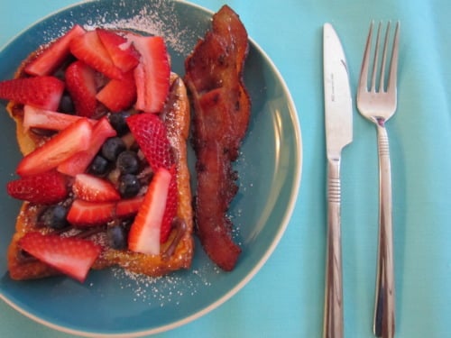 French toast topped with fresh strawberries and blueberries, served with a slice of crispy bacon on a blue plate.