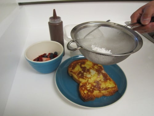French toast on a blue plate being dusted with powdered sugar through a fine mesh strainer, served with a bowl of fresh berries and a bottle of chocolate sauce.