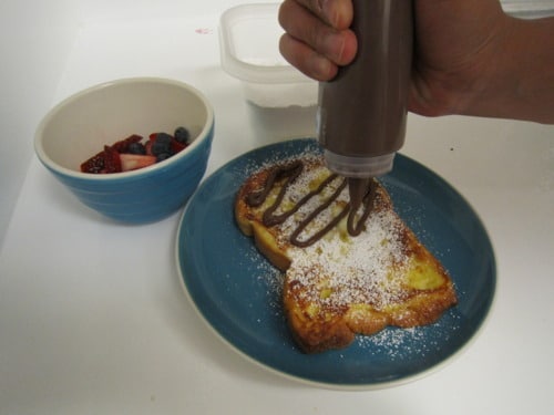 French toast dusted with powdered sugar being drizzled with chocolate sauce, served with a side bowl of fresh mixed berries.