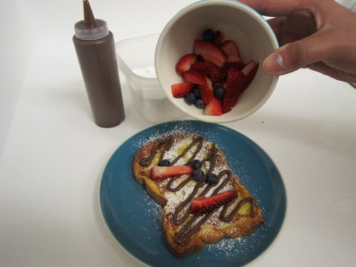 French toast topped with powdered sugar and chocolate drizzle on a blue plate, with fresh berries being added from a small bowl.