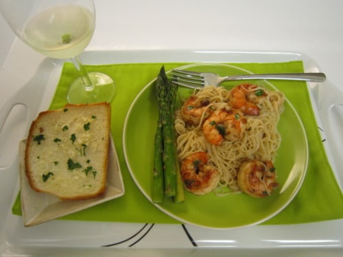 Plate of shrimp scampi with angel hair pasta, served with roasted asparagus, garlic bread, and a glass of white wine on a green placemat.