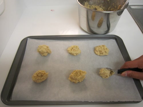 Cookie dough scooped onto a parchment-lined baking sheet, with a hand using a spoon to portion dough for white chocolate butterscotch chip cookies.