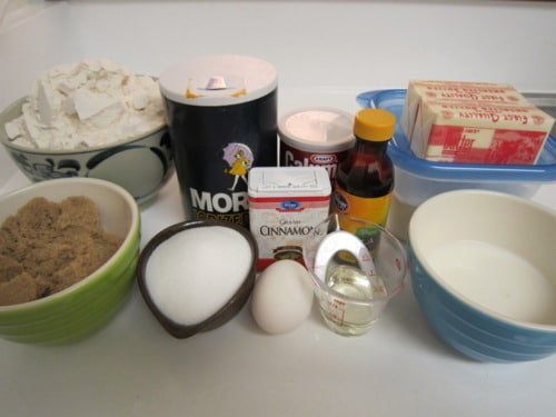 Ingredients for crumb cake, including flour, salt, baking powder, brown sugar, granulated sugar, cinnamon, egg, oil, milk, vanilla extract, and butter, arranged on a white surface.