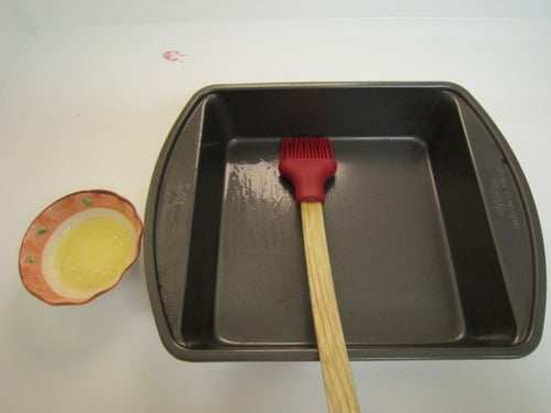 Square baking pan being brushed with melted butter using a red silicone brush, prepared for making crumb cake.