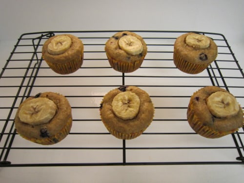 Banana blueberry muffins topped with banana slices cooling on a black wire rack.