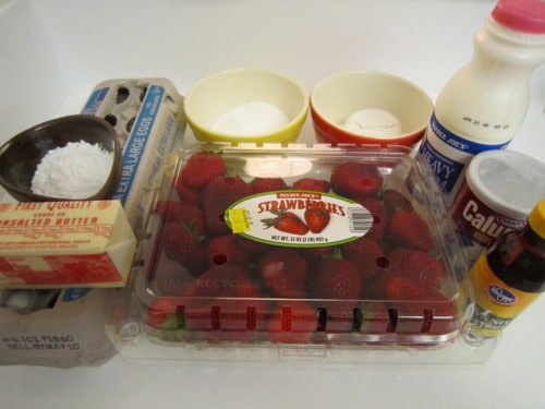 Ingredients for strawberry torte, including fresh strawberries, butter, eggs, sugar, powdered sugar, flour, heavy cream, and vanilla extract, arranged on a countertop.