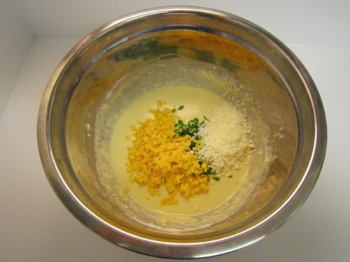 Batter in a mixing bowl with shredded cheddar cheese, grated Parmesan, and chopped chives, prepared for cheesy popovers.