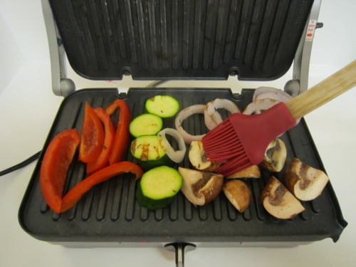 Sliced red bell peppers, zucchini, onions, and mushrooms being brushed with oil while grilling on an indoor electric grill for herb grilled vegetables.