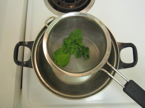 Blanched spinach leaves in a mesh strainer over a pot on the stove, prepared for pan-seared scallops with Yukon gold potato mash.