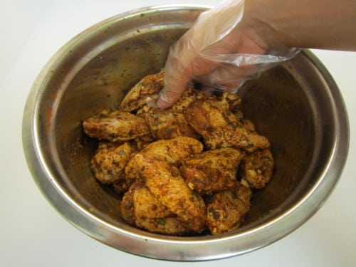 Gloved hand mixing seasoned chicken wings in a stainless steel bowl