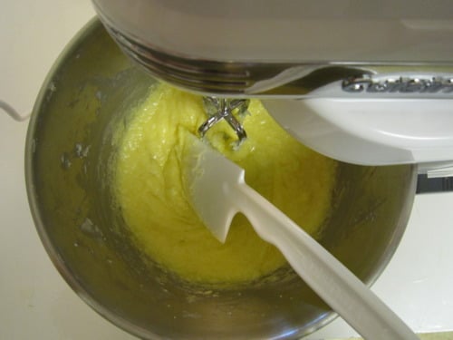 Yellow cupcake batter being mixed in a stand mixer with a spatula resting in the bowl, prepared for orange buttercream cupcake cones.