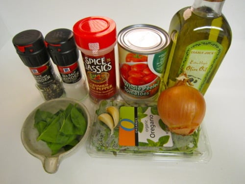 Ingredients laid out for cooking, including basil leaves, oregano, garlic cloves, a yellow onion, canned whole tomatoes, olive oil, black pepper, crushed red pepper, and seasoning containers.