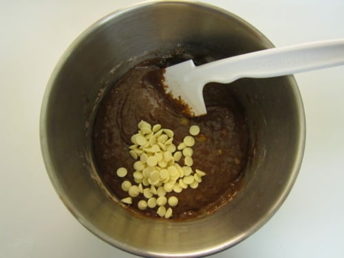 White chocolate chips added to chocolate banana muffin batter in a mixing bowl with spatula.