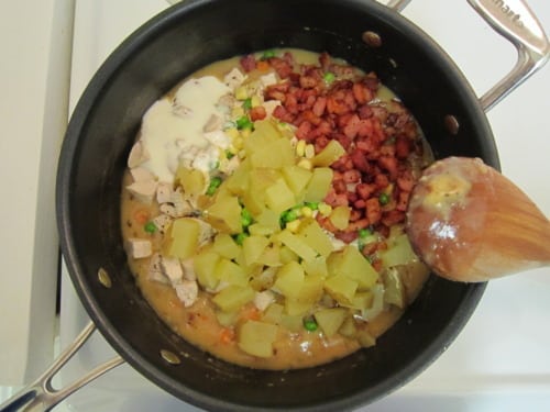 Diced turkey, pancetta, potatoes, mixed vegetables, and cream in a skillet being stirred with a wooden spoon, prepared for turkey and pancetta pot pies.