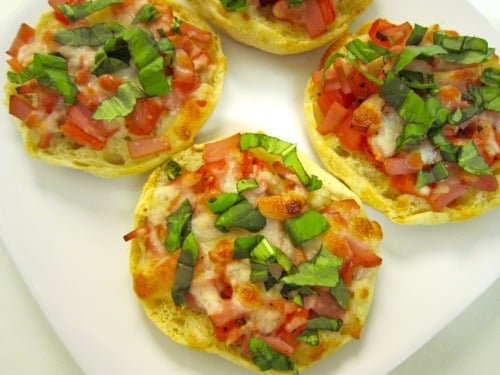 Four mini English-muffin pizzas topped with melted cheese, chopped tomatoes, and fresh basil.