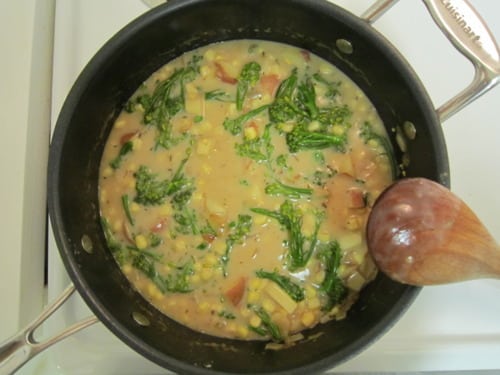 Broccoli chowder with corn and bacon simmering in a saucepan, stirred with a wooden spoon.