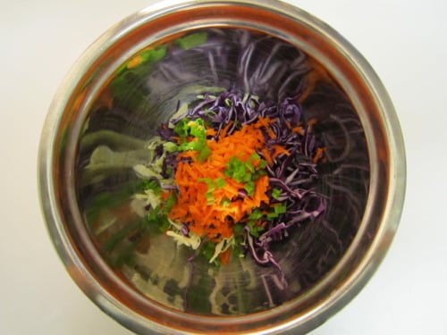 Shredded red cabbage, carrots, green onions, and cilantro in a stainless steel mixing bowl, prepared for beer-battered fried trout tacos with spicy horseradish.