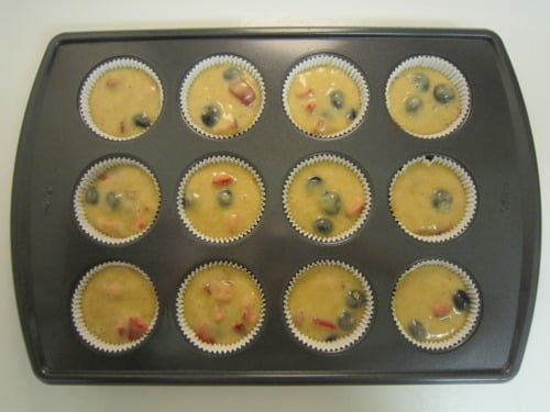 Muffin tray filled with paper liners and uncooked batter containing strawberries, blueberries, and banana for lemon fruit muffins.