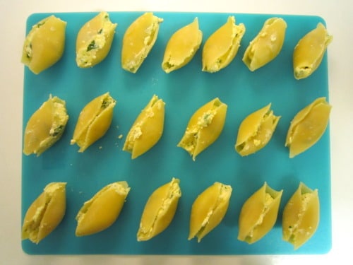 Uncooked pasta shells filled with ricotta cheese mixture arranged on a blue cutting board, prepared for stuffed shells with arrabbiata sauce.