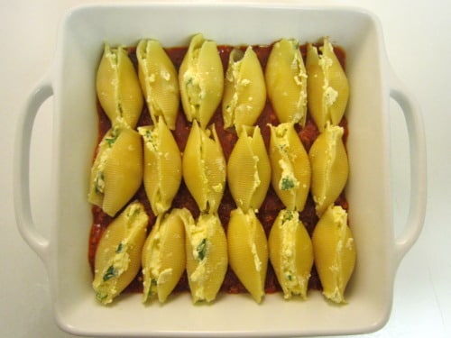 Unbaked stuffed pasta shells arranged neatly in rows in a white square baking dish, filled with a ricotta and herb mixture on top of a layer of red arrabbiata sauce.