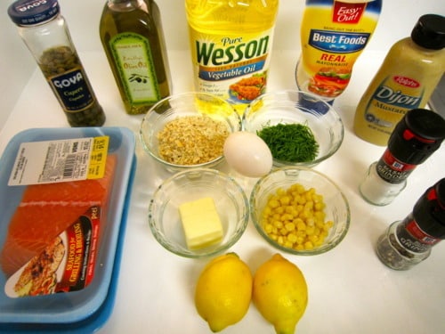 Ingredients for salmon cakes arranged on a white countertop, including fresh salmon, breadcrumbs, egg, corn, herbs, lemon, butter, oil, and assorted seasonings.