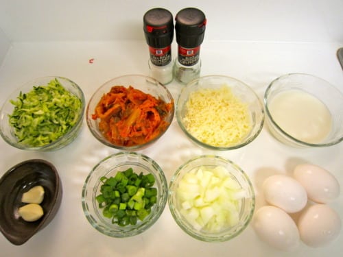 Ingredients for zucchini and kimchi quiche, including shredded zucchini, kimchi, cheese, milk, eggs, chopped onions, green onions, garlic, salt, and pepper.