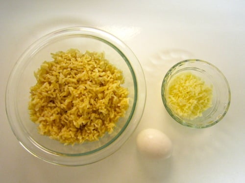 Bowls of cooked brown rice and shredded cheese with a whole egg on a white surface, prepped for cooking
