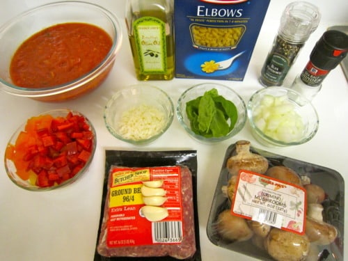 Ingredients for Italian goulash, including ground beef, pasta, tomato sauce, mushrooms, garlic, onions, bell peppers, spinach, cheese, olive oil, and seasonings, arranged on a counter.