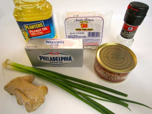 Ingredients for crab and cream cheese wontons, including peanut oil, wonton wrappers, sea salt, canned crab meat, cream cheese, green onions, and fresh ginger, arranged on a white surface.