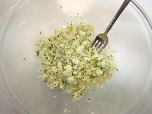 Glass bowl with crumbled cheese mixed with herbs, stirred using a fork.