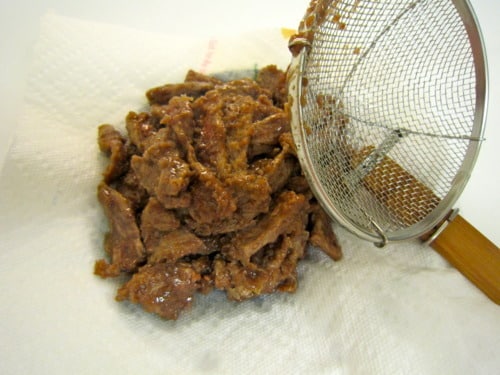 Fried strips of beef drained on a paper towel with a wire skimmer, prepared for Mongolian beef.