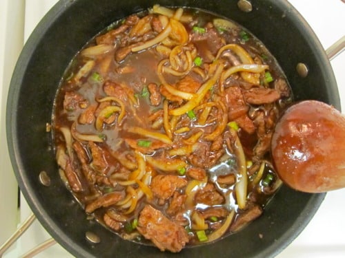 Mongolian beef simmering in a saucepan with sliced onions and green onions in a rich, dark sauce.