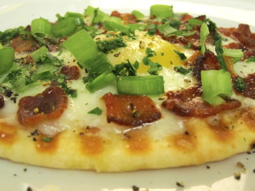 Grilled breakfast pizza topped with crispy bacon, a baked egg, melted cheese, and fresh chopped green onions and herbs.