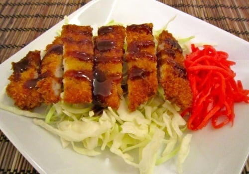 Tonkatsu pork cutlet sliced and served over shredded cabbage with sauce drizzle and a side of pickled ginger on a white plate.