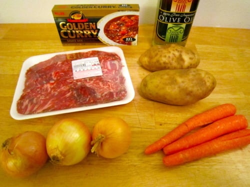 Ingredients for Korean curry laid out on a wooden surface, including beef, potatoes, carrots, onions, curry sauce mix, and olive oil.