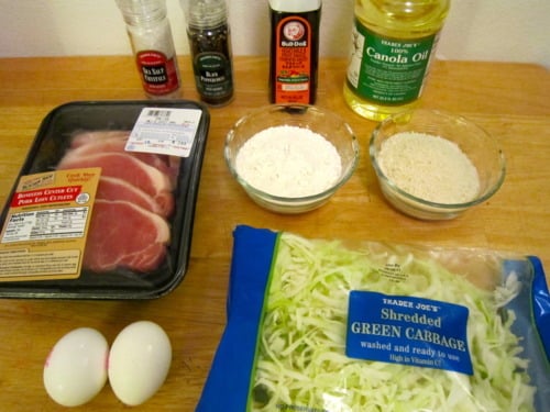 Ingredients for tonkatsu pork cutlet, including pork loin chops, eggs, flour, panko breadcrumbs, shredded green cabbage, canola oil, soy sauce, black pepper, salt, and tonkatsu sauce, arranged on a wooden surface.