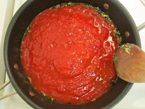 Tomato sauce simmering in a black saucepan with garlic and herbs, being stirred with a wooden spoon for meatball subs.