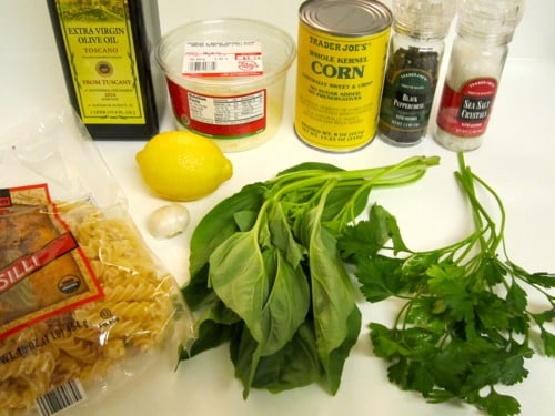 Ingredients for sweet corn pasta including fusilli, fresh basil, parsley, lemon, garlic, canned corn, Parmesan cheese, olive oil, salt, and pepper.