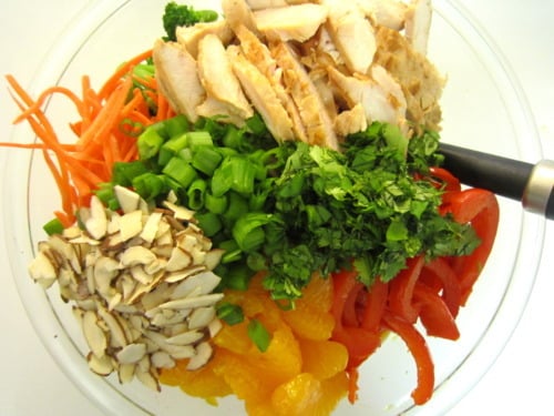 Ingredients for Asian chicken noodle salad in a glass bowl, including sliced grilled chicken, red bell peppers, mandarin oranges, sliced almonds, green onions, shredded carrots, cilantro, and broccoli.