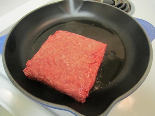 Block of raw ground beef in a black skillet on the stovetop, beginning to cook for sour cream noodle bake.