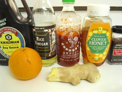 Ingredients for spicy citrus sauce, including soy sauce, rice vinegar, sriracha, honey, red pepper flakes, orange, and fresh ginger, arranged on a countertop.