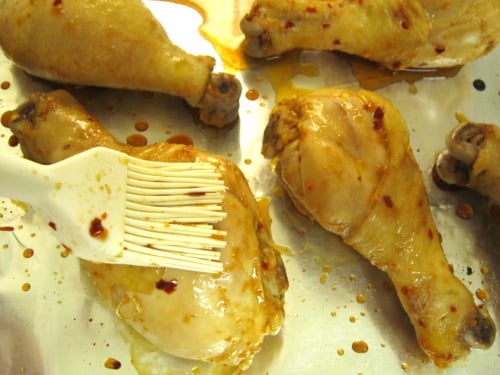 Chicken drumsticks being brushed with a spicy citrus glaze on a baking sheet, prepared for steaming and serving with cilantro dipping sauce.
