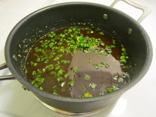 Sauce with chopped green onions simmering in a black saucepan on a stovetop.