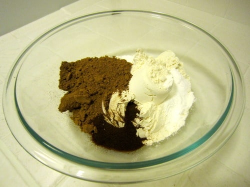 Glass mixing bowl with dry ingredients for double chocolate muffins, including flour, cocoa powder, and espresso powder.