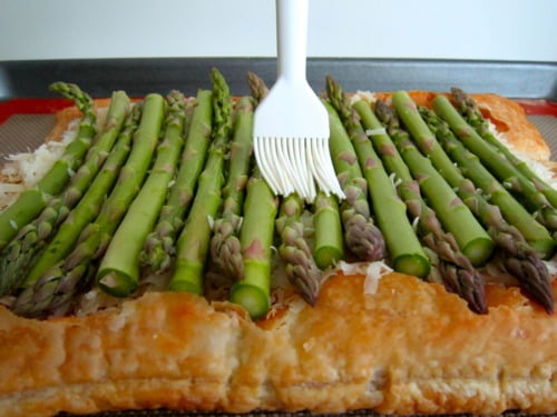 Fresh asparagus being brushed with oil on top of a golden puff pastry tart layered with shredded Gruyère cheese.