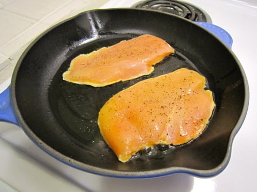 Two raw chicken breasts seasoned with pepper cooking in a black skillet.