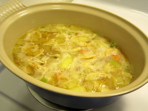 A pot of chicken noodle soup simmering on the stove, filled with broth, shredded chicken, noodles, and diced vegetables.
