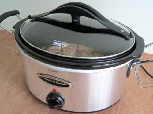Slow cooker with a glass lid partially showing meat inside, in the process of cooking chicken adobo.