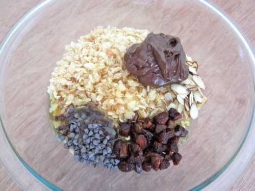 Glass bowl with ingredients for crunchy Nutella granola bars, including oats, Nutella, sliced almonds, chocolate chips, raisins, and crushed cereal.