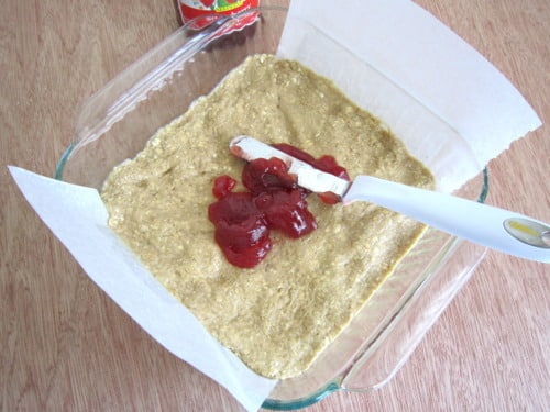 Strawberry jam being spread over a layer of oat mixture in a parchment-lined glass baking dish, prepared for strawberry honey oatmeal bars.